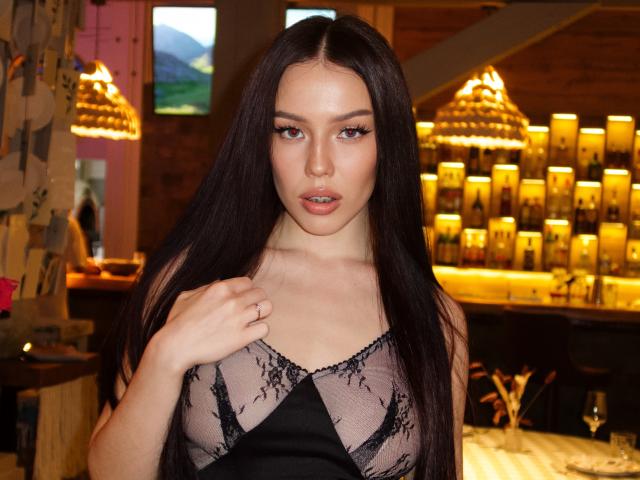 JaneWow with long dark hair, wearing a sheer black lace top, has her hand on her chest, showing off her tits, in a warmly lit bar.