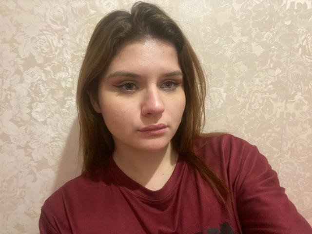 Heike Rippy, con i capelli castani, indossa una maglietta rossa, ha un'espressione seria, un seno piccolo e un trucco leggero con un sottile filo di eyeliner. Lo sfondo è una parete con un motivo floreale..