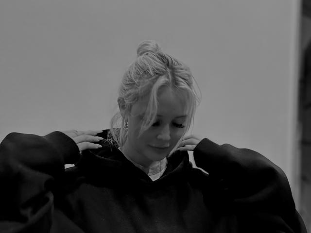 AnnamariaVillarin, black-and-white photo, blonde hair in a messy bun, wearing a dark hoodie, eyes down, hands adjusting hood, slight smile, soft light, subtle shadows, calm expression.