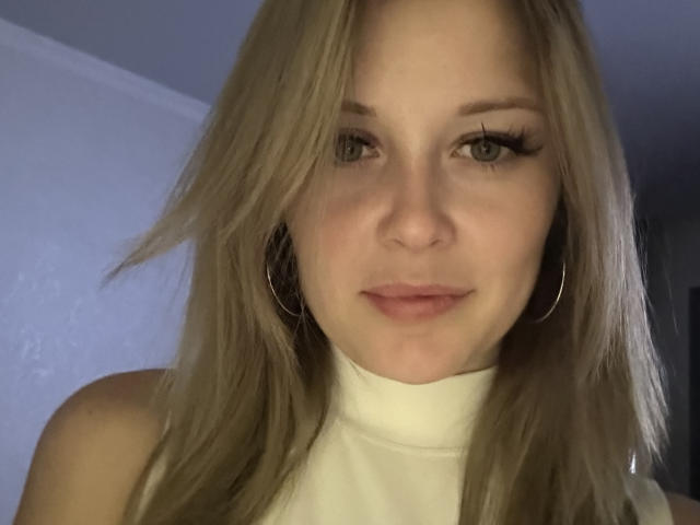 AnnamariaVillarin has blonde hair, green eyes, and is wearing a white turtleneck with hoop earrings, looking at the camera with a slight smile. Her tits are not visible.