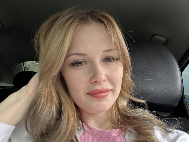 AnnamariaVillarin has blonde hair, green eyes, and pink lipstick, wearing a white shirt over a pink top, with a hand in her hair, smiling in a car.