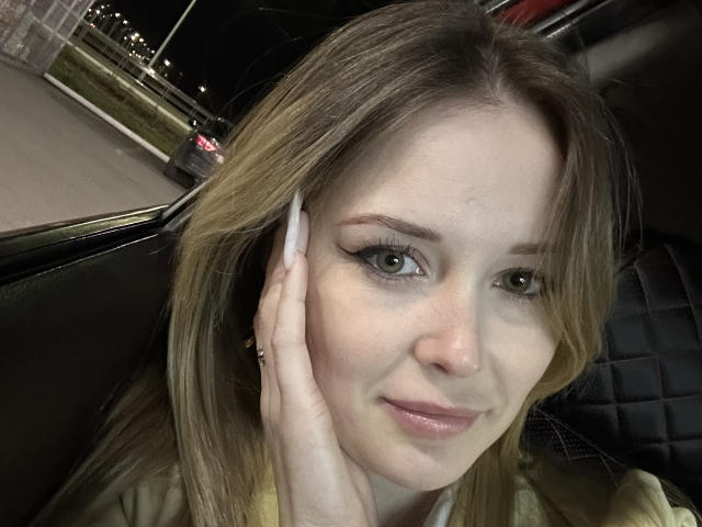 AnnamariaVillarin has light brown hair, green eyes, and long white nails, touching her face with a slight smile, wearing a yellow top, in a car at night.