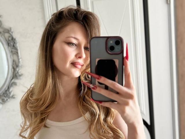 AnnamariaVillarin with wavy blonde hair, wearing a white tank top, holding a phone with red nails, taking a selfie, smiling slightly, in a well-lit room.