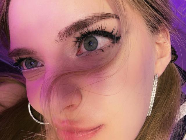 EdrisLueder's face with green eyes, black eyeliner, long lashes, and pinkish skin. She's wearing a silver hoop earring and has light brown hair, slightly covering her left eye.