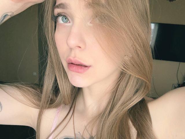 EdrisLueder has light blue eyes, pink lips, and long brown hair with a side part, showing a tattoo on her arm, and wearing a pink top, looking into the camera.