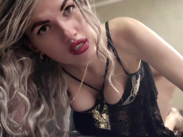 MilaLoona with wavy blonde hair, red lips, and black lace lingerie, leaning forward, showing her tits, a gold chain necklace, and a slightly open mouth.