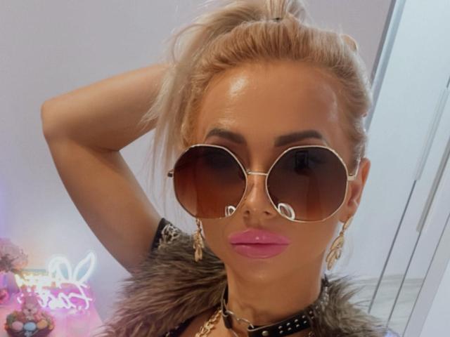 MoniqueBliss with shiny tan skin, pink lips, blonde ponytail, wearing oversized brown sunglasses, gold earrings, fur collar, and black studded choker, looking sexy and confident.