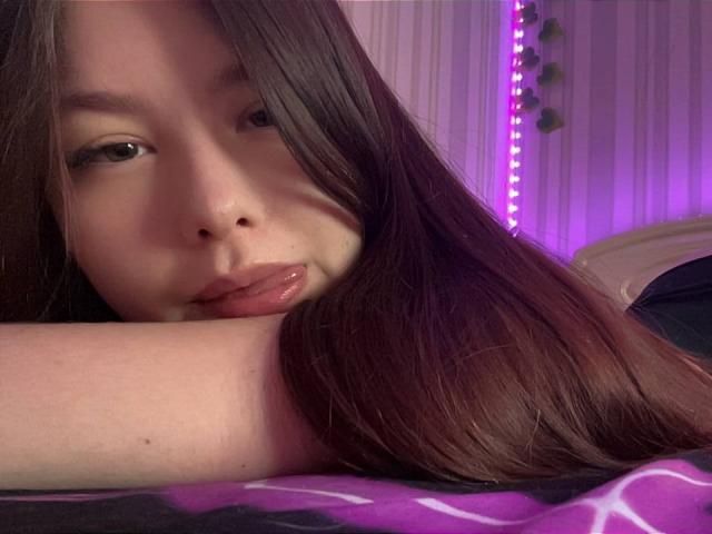 KandraPutney with long brown hair, pouty lips, and a cute face, lying down with her arm over her forehead, looking at the camera with a slight smile.