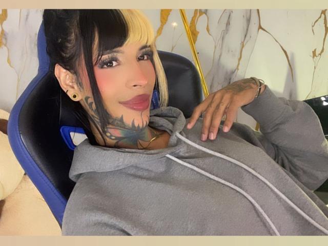 AnneKisss, con tatuaggi sul collo e sulla mano, capelli biondi e neri, indossa una felpa grigia, è seduta su una sedia blu, sorride, mostrando il rossetto rosa e la gola tatuata..