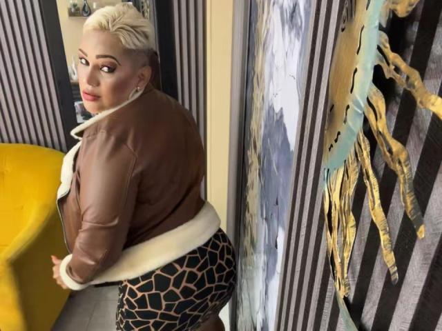 GianellaCox with short blonde hair, brown leather jacket, white fur collar, and black leopard print pants, looking over her shoulder, standing next to a yellow chair.