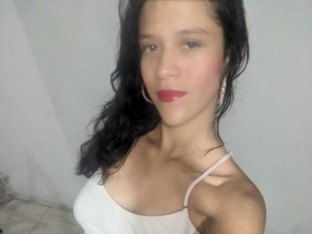 LunaWarner with black wavy hair, red lipstick, white spaghetti strap top, showing her tits and earrings, light skin, slightly flushed cheeks, and a neutral expression.