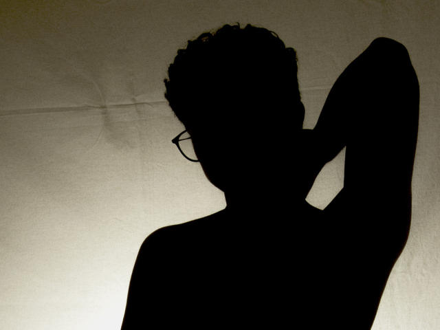 AdoniBaskerville in controluce, con capelli ricci, braccio sollevato e occhiali, una figura scura in contrasto con una parete chiara e strutturata, che mette in risalto la parte superiore del corpo muscolosa..