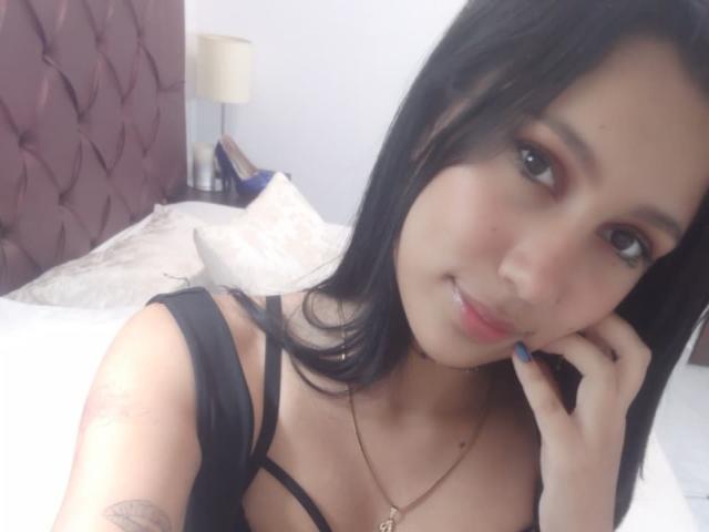 KataleyaMansson with black hair, light skin, and a small tattoo on her left arm, wearing a black bra, resting her chin on her hand, smiling at the camera.