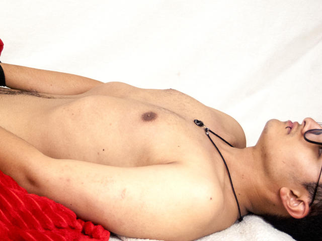 AdoniBaskerville est allongé sur une surface blanche recouverte d'un tissu rouge, torse nu, montrant de petits tétons bruns et un collier noir avec un pendentif. Il porte des lunettes et a les cheveux noirs..