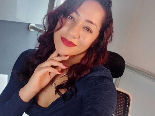 SamanthaCorine with wavy dark red hair, wearing a navy blue top, red lipstick, and red nail polish, touching her chin with a gentle smile. Boobs visible under top.