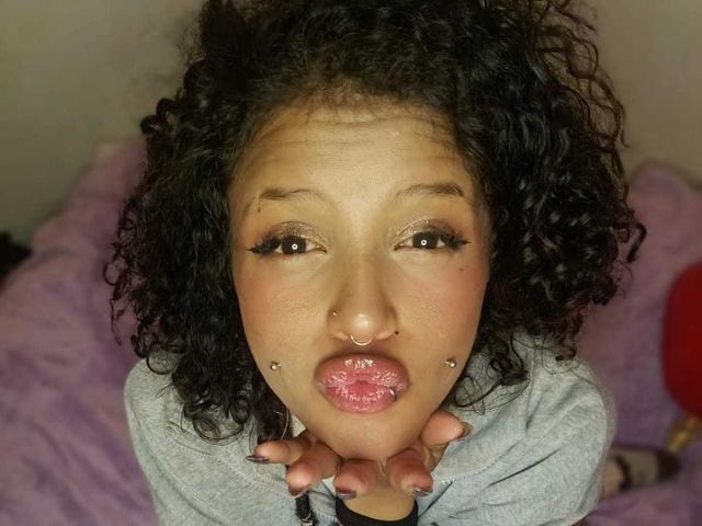 LunaKeller has curly black hair, brown eyes with thick eyelashes, a nose ring, lip piercings, and a finger near her shiny, wet pink lips. She wears a gray shirt.