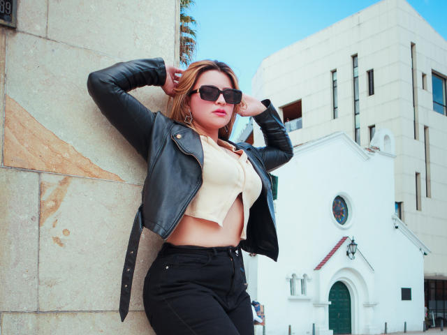 SaraWestt with long brown hair, pink lipstick, white strapless top, touching her pink-manicured fingers to her mouth, tattoo of an ankh on her right shoulder.