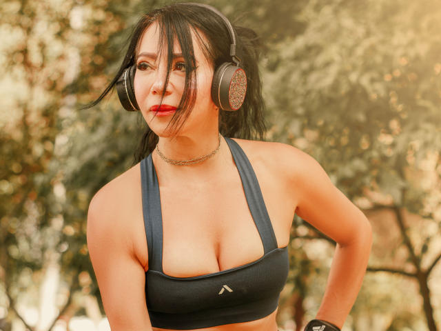HelenHard with medium tits, wearing black sports bra, headphones, red lipstick, black choker, and bracelet, standing outdoors, looking to the side, sunlight on her face.