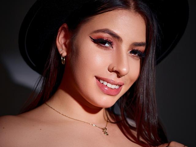 VanessaClose has shiny brown hair, a black hat, and a nose ring. She's smiling with pink lipstick, dark eyeliner, and a gold necklace. Her tits are barely visible.