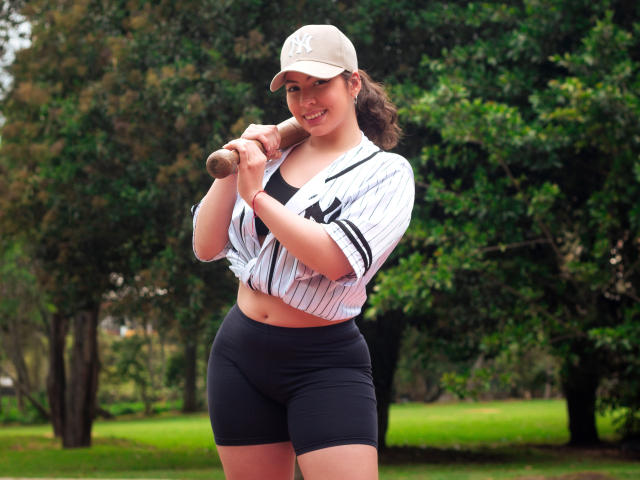VanessaClose with curvy tits, wearing a baseball cap and pinstripe jersey, holding a bat, black shorts, smiling, standing in a green outdoor setting, her ass slightly visible.
