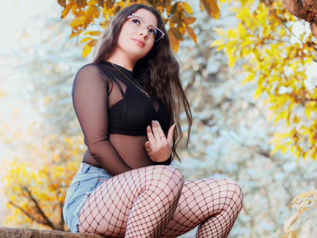 VanessaClose with long brown hair, glasses, black sheer top, denim shorts, fishnet stockings, sitting outdoors, hands on chest, yellow autumn leaves in background, confident, sexy.