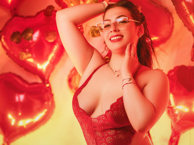 VanessaClose with glasses, red lace lingerie, smiling, hands up, showing her tits, pink balloons in background, red lighting, looking sexy, wearing bracelets, smiling, red lipstick.