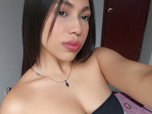 Angels69v has medium brown skin, black straight hair, and pink lipstick. She's wearing a black strap top, showing her bare shoulders and small, firm tits. Necklace with purple charm.