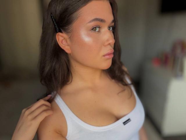 BerryZegarelli with shiny skin, brown hair in a clip, wearing a white tank top showing her tits, looking to the side with a pouty face, in a blurry background.