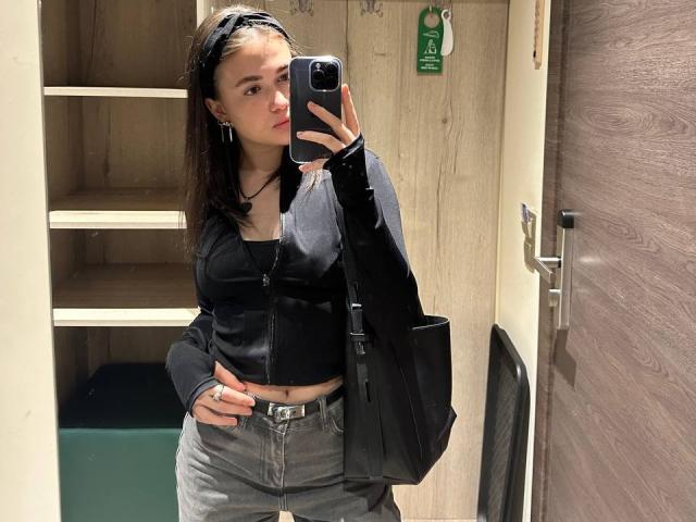 BerryZegarelli with black headband, black zip-up top, gray jeans, black bag, and ring, taking mirror selfie, showing pouty face, holding phone, in dimly lit room.
