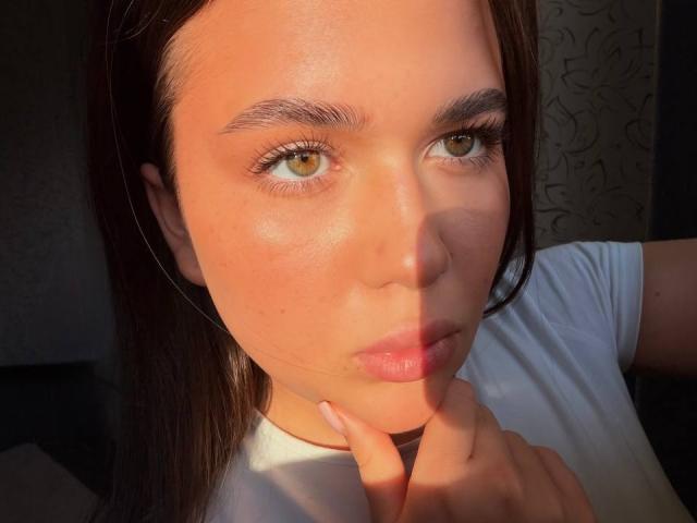 BerryZegarelli's face with warm sunlight, brown eyes, full pink lips, straight dark brown hair, white shirt, hand touching neck, slightly flushed skin, subtle makeup, looking into camera.