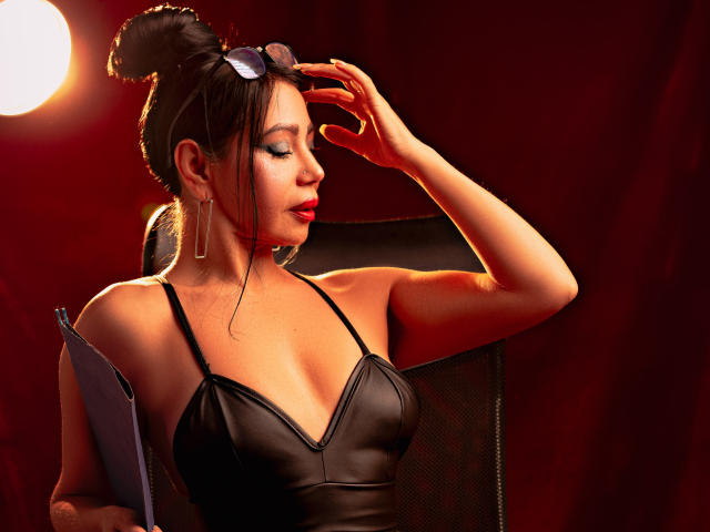 HelenHard with dark hair in a bun, wearing a black leather top, touching her head with sunglasses on. Her tits are visible, holding a clipboard. Red background.