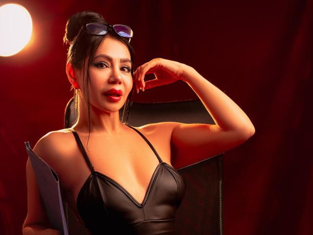 HelenHard with black leather top, red lipstick, dark hair in bun, sunglasses on head, holding clipboard, arm resting on chair, warm red light, medium shot, sexy, confident.