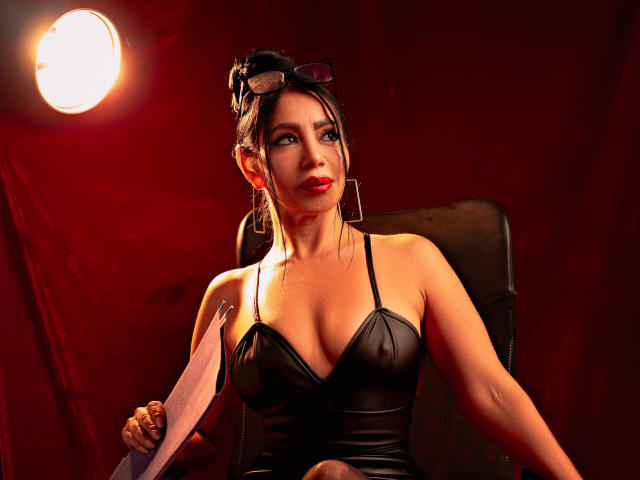 HelenHard with dark hair in a bun, wearing a black leather top, holding papers, sitting in a chair, red light, sunglasses on head, looking to the side, red background.