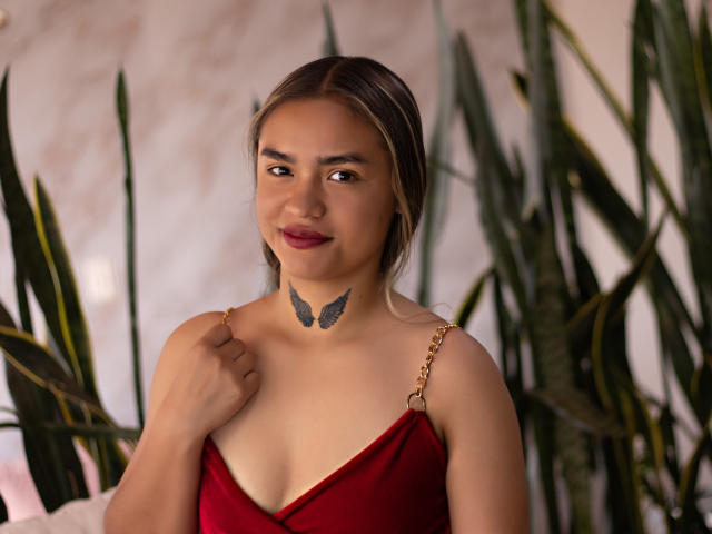 MollyRed with brown eyes, red dress, and butterfly tattoo on her neck, holding dress strap, smiling, surrounded by green plants. Boobs visible, medium-sized, cute, and sexy.