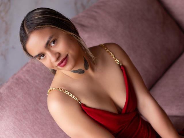 Molly, pele clara e morena, vestido vermelho, correntes de ouro, seios pequenos, tatuagem preta no pescoço e mechas loiras, sentada em um sofá rosa, sorrindo, olhando para cima..