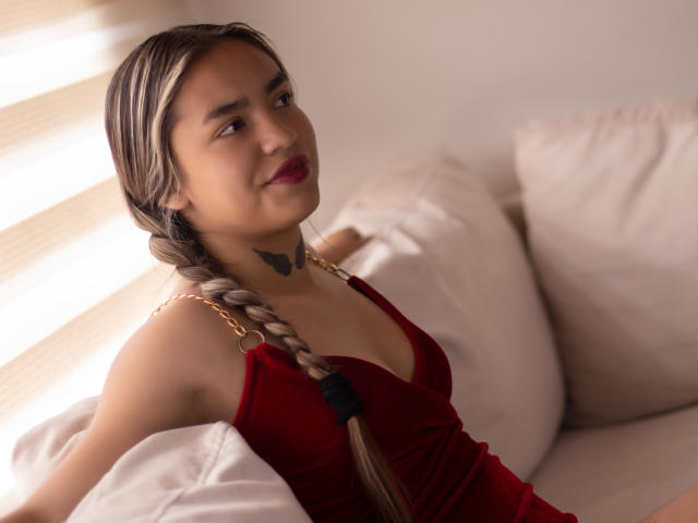AnnaLadera with blonde braids, red velvet dress, and butterfly tattoo, sitting on white cushions, smiling, sunlight on her shoulder. Her tits are small, and she has red lipstick.