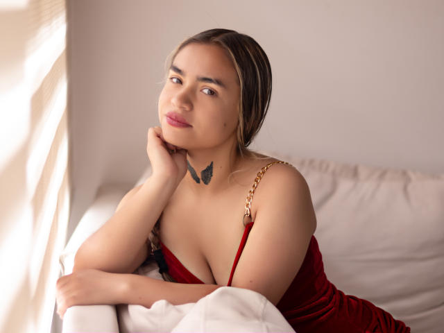 AnnaLadera with light brown hair, red dress, and butterfly tattoo, resting chin on hand, showing her tits, soft smile, on white bed, sunlight on left.