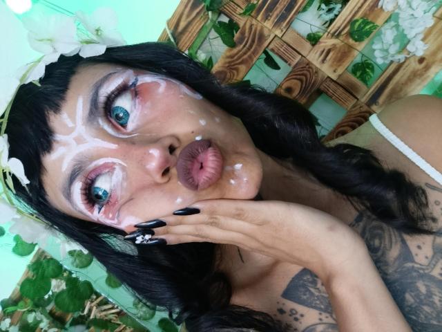 NyxVega has cum on her face, pouty pink lips, blue eyes, black nails, white flower headband, and tattoos, with long dark hair and a green background.