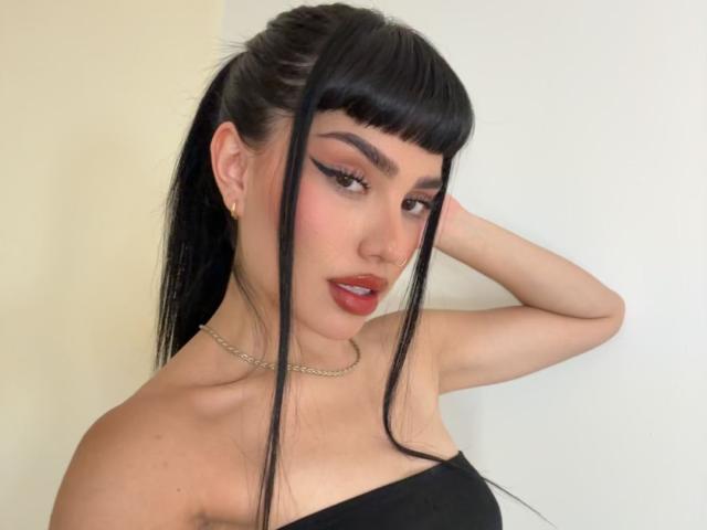 Almaa, met lang zwart haar in een hoge paardenstaart, pony en gevleugelde eyeliner, draagt een zwarte top zonder bandjes, een choker en rode lipstick, met één hand op haar hoofd..