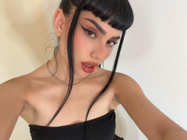 AlmaaBlaze, con coletas negras, delineador de ojos marcado y lápiz labial rojo, vestía un top negro sin tirantes, un collar de cadena dorada y aretes dorados, mostrando sus pequeños senos y una ligera escote..