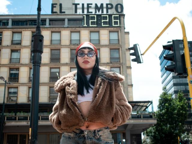 Almaa, con un gorro rojo, gafas de sol oscuras, una chaqueta marrón y suave, un top corto blanco y jeans de cintura alta, está de pie frente a un edificio, con semáforos al fondo..