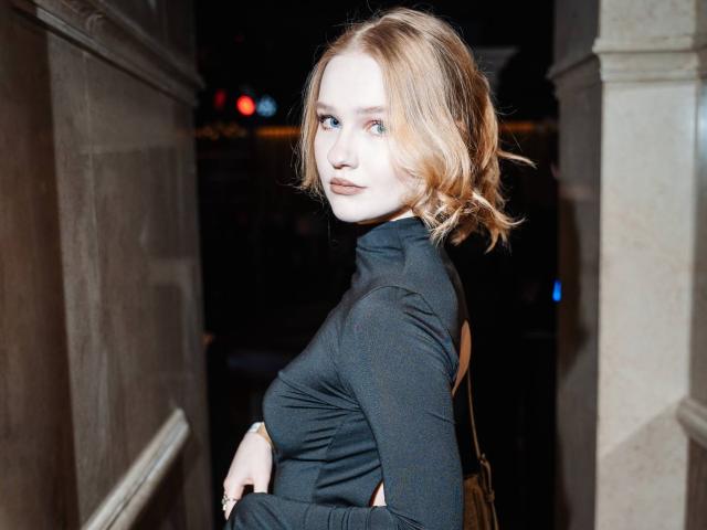 OlympiaCheroki with short, wavy blonde hair, blue eyes, and a serious look, wearing a black, high-neck, long-sleeve top, standing in a dimly lit, narrow hallway.