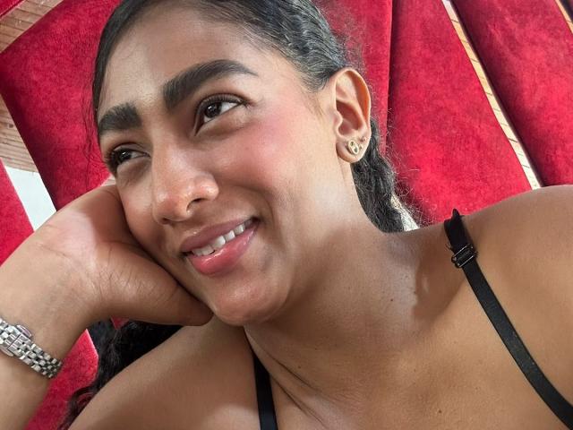SamanthaVenus, desnuda, con la piel húmeda, senos grandes y un tatuaje en el hombro derecho, está recostada en una bañera con azulejos oscuros, con las piernas separadas y la mano derecha tocándose la cara, de labios rojos..