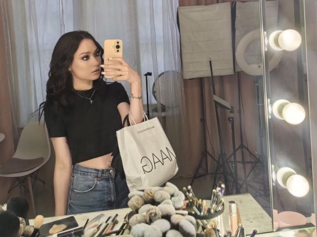 PatienceObremski with long dark hair, black crop top, blue jeans, holding a white shopping bag, mirror selfie, surrounded by makeup, ring lights, and a chair.