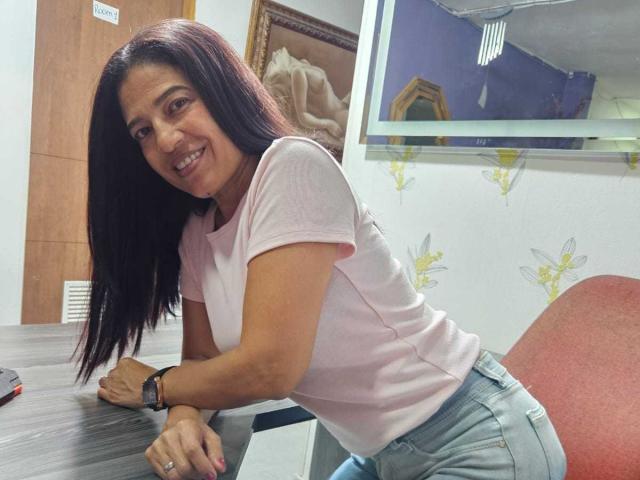 Fresyta, with medium tits and dark brown hair, leans on a table in a pink shirt and blue jeans, smiling at the camera. She's wearing a black wristwatch.