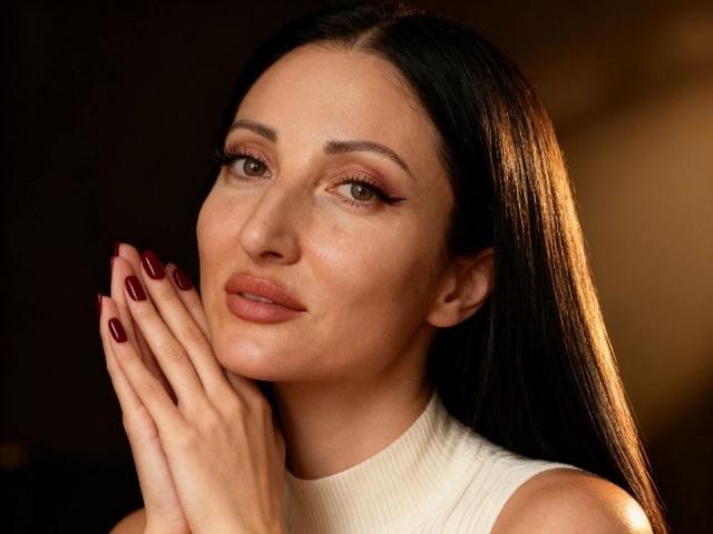 MissRossi with long black hair, brown eyes, and red lipstick, wearing a white sleeveless top, hands together with red nails, slightly smiling, in a warm-lit portrait.