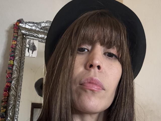 SeleneBrigth with brown bangs, wearing a black hat, pink glossy lips, and a slightly pouty expression, her nose and mouth prominent, in a selfie-style photo.