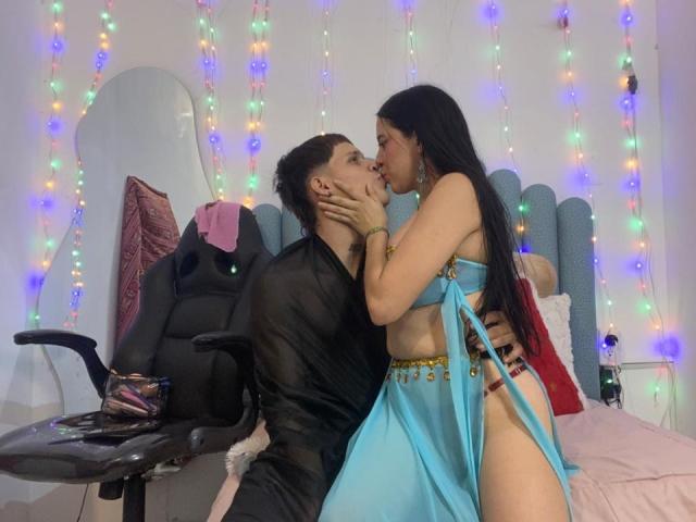 AnjaliRohan in a blue harem-style outfit with gold accents, kissing a guy in a black sheer shirt, holding his face, in a bedroom with colorful fairy lights.