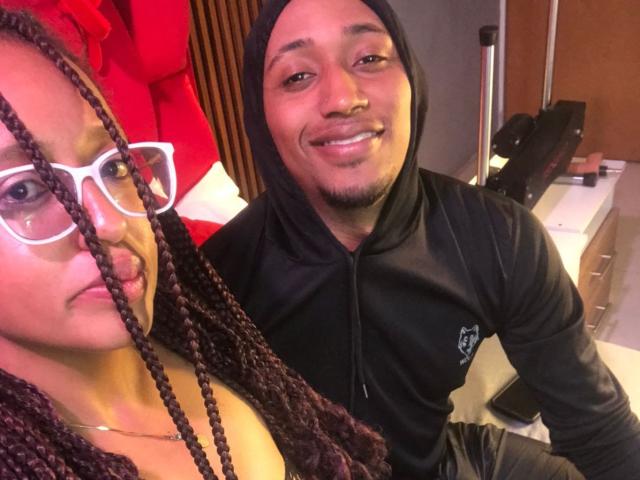 LilithAndAsmodeo with braided hair, white glasses, and visible tits, sits next to LilithAndAsmodeo in a black hoodie, smiling, in a well-lit room.