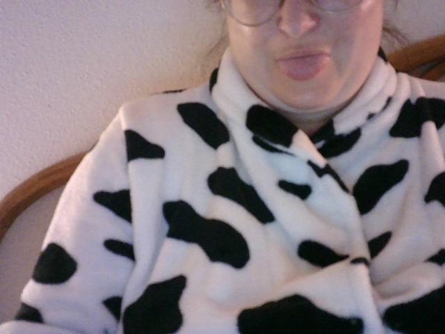 DominaBlonddi wearing a black-and-white cow pattern robe, pouting with her lips, glasses on, slightly chubby face, visible chin and neck, blurry photo.